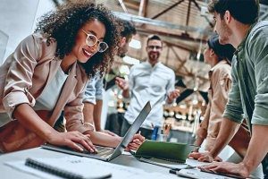 A group of people talking to one another and one of them is working on a laptop.